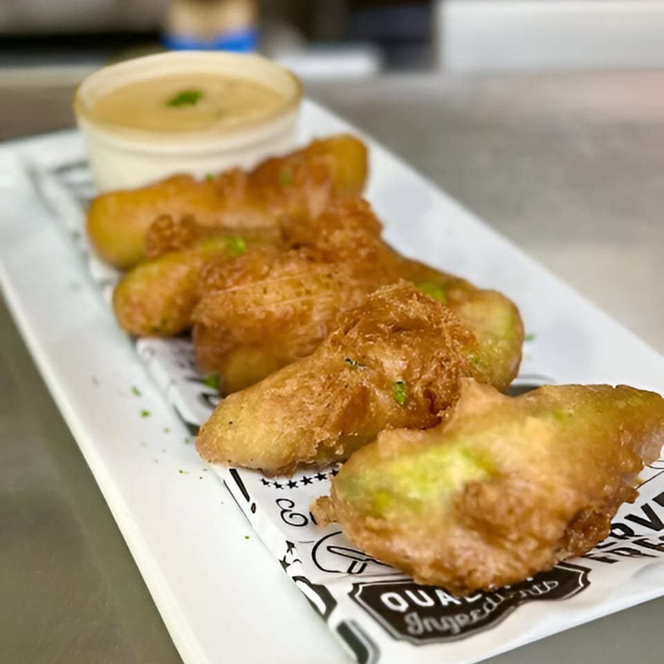 Golden-brown fried pickles with a side of creamy dipping sauce on a white rectangular plate.