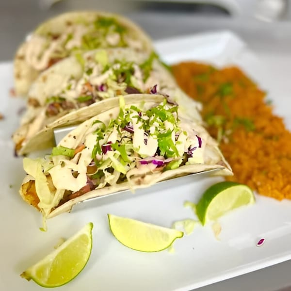 Three fish tacos with shredded cabbage, sauce, and lime wedges, served with orange rice.