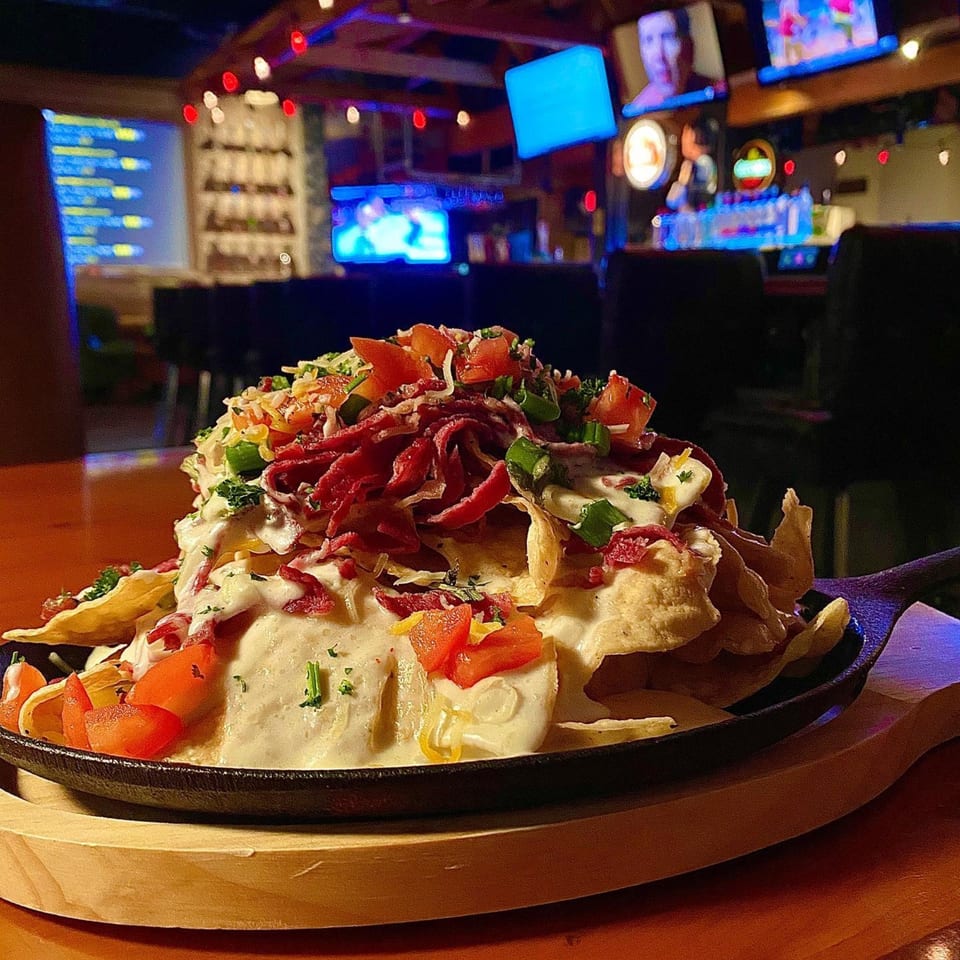 Loaded nachos with corned beef, cheese sauce, tomatoes, and green onions in a sports bar setting.