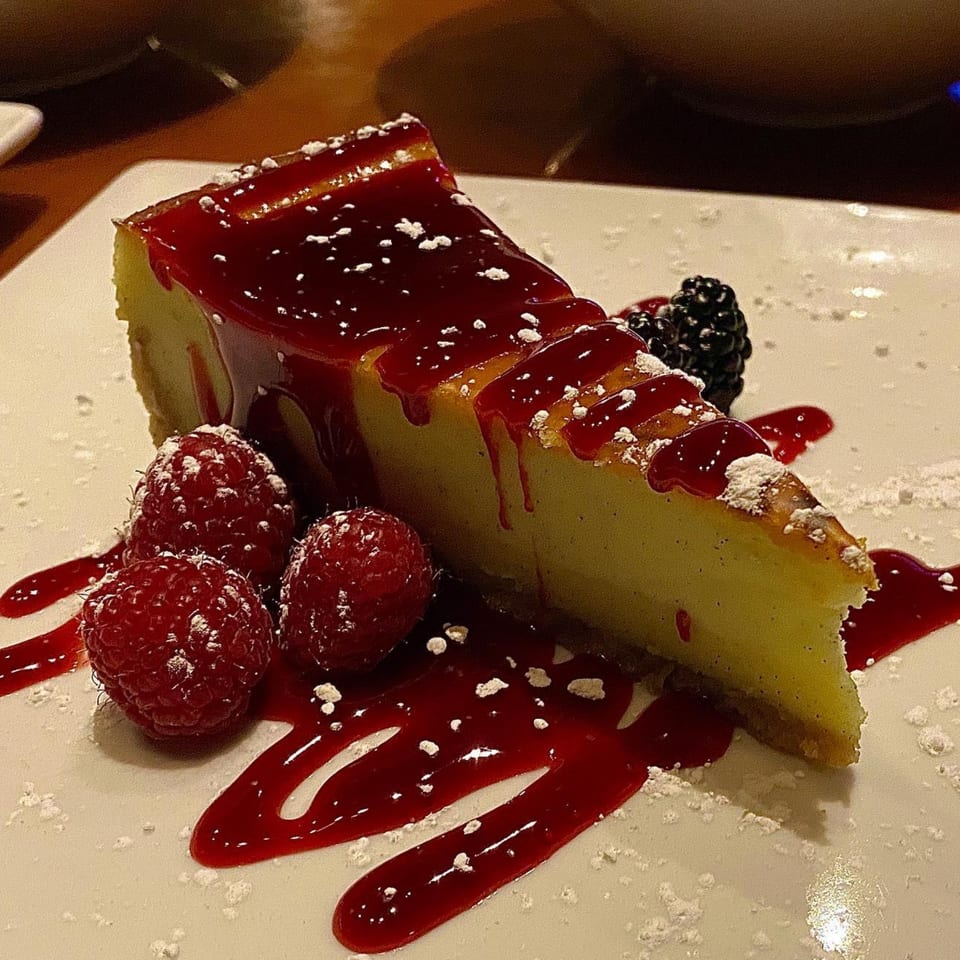 A slice of cheesecake with raspberry sauce, fresh raspberries, and a blackberry on a white plate.