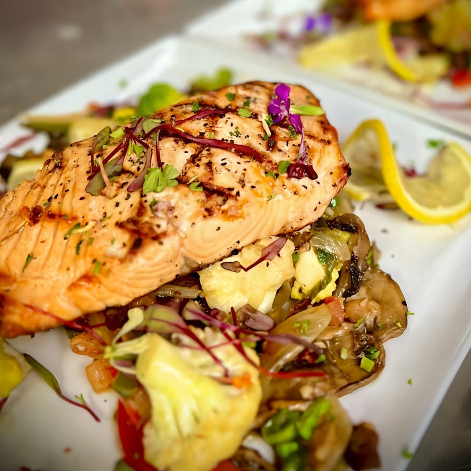 Grilled salmon with herbs and a purple edible flower, served over sautéed vegetables and lemon.