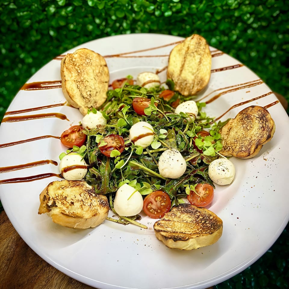 A fresh salad with greens, cherry tomatoes, mozzarella balls, and toasted bread on a white plate.