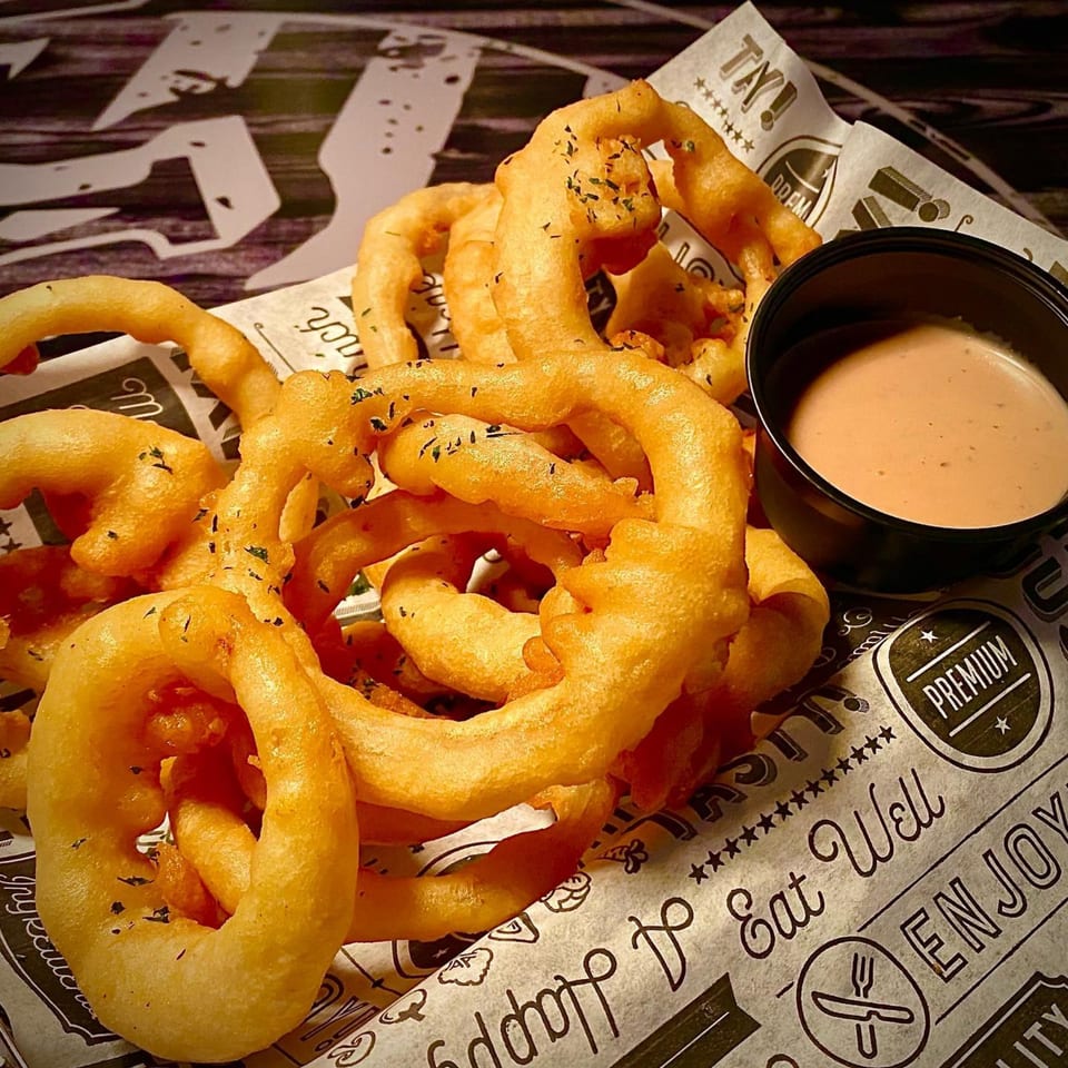 A basket of golden curly fries and onion rings with a side of dipping sauce at Joey's Tavern.